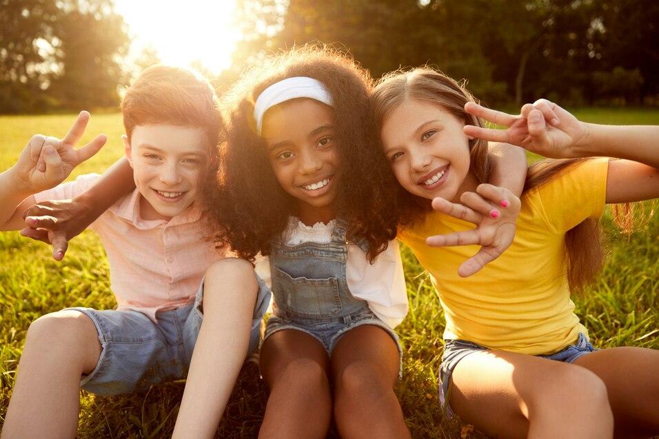 Drei fröhliche Kinder sitzen auf der Wiese und zeigen das Peace-Zeichen, umgeben von Sonnenlicht und Grün.